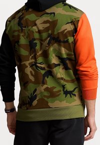 Mężczyzna w bluzie z kapturem w kamuflażowym wzorze, z jedną czarną rękawą i jedną pomarańczową, pokazany od tyłu.