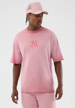 Uomo che indossa un cappellino rosa dei New York Yankees, una T-shirt rosa coordinata e pantaloni, che guarda verso il basso a sinistra su uno sfondo semplice.
