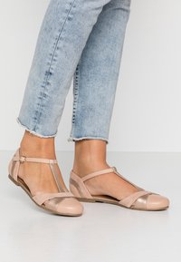 Pies usando zapatos planos de tira en color beige, combinados con unos vaqueros cortos de dobladillo deshilachado en color azul claro, de pie sobre un suelo blanco.