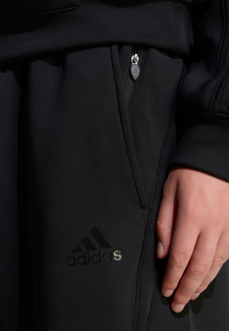 Schwarzer sportlicher Jogger mit seitlicher Reißverschlusstasche, glatter Stoffstruktur und reflektierendem Adidas-Logo nahe dem Saum. Hand ruht auf dem Stoff.