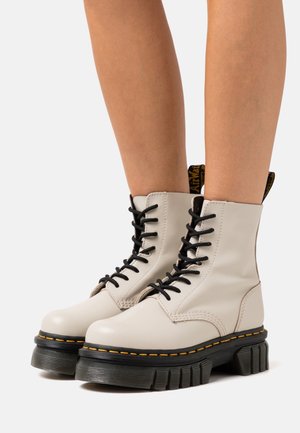 Bottes montantes en cuir crème avec lacets, dotées d'une semelle en caoutchouc noir épaisse et de surpiqûres jaunes. Elles comportent un laçage noir à l'avant et une languette.