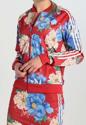 Femme portant une veste rouge Adidas et une jupe avec un grand imprimé floral bleu et blanc et des rayures blanches sur les manches et les côtés de la jupe.