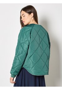 Femme aux longs cheveux bruns portant une veste matelassée turquoise et une jupe bleu marine, regardant par-dessus son épaule sur un fond uni.