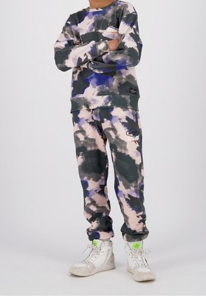 Persona vistiendo sudadera y pantalones deportivos a juego con diseño tie-dye en tonos gris, negro, púrpura y beige, con zapatillas altas blancas.