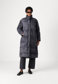 Juodas pamuštas ilgas paltas su aukštu apykakle, turintis horizontalius quilted skyrius ir šonines kišenes, dėvimas virš plačių tamsių džinsų ir slip-on batų.
