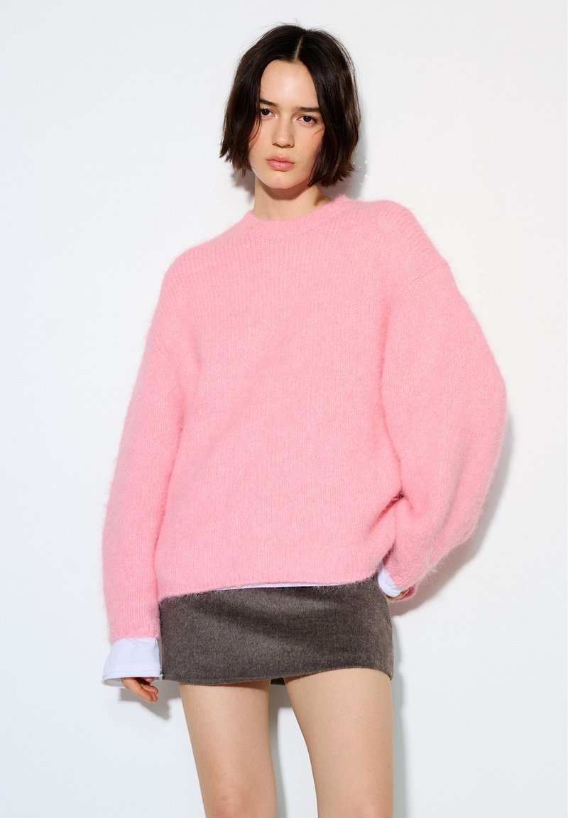 Jeune femme portant un pull rose ample et une jupe courte gris foncé, debout devant un fond blanc.