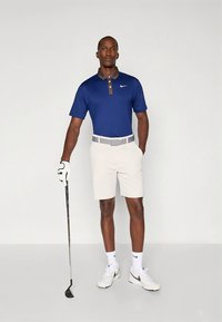 Tumši zila polo krekls ar brūnu apkakli, gaiši bēšas šorti, baltas zeķes un Nike sporta apavi. Tur rokā golfa nūju, stāv uz vienkārša fona.