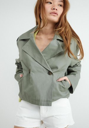 Młoda kobieta z długimi włosami nosi luźną zieloną marynarkę dwurzędową i białe postrzępione dżinsowe szorty, ręce w kieszeniach marynarki.