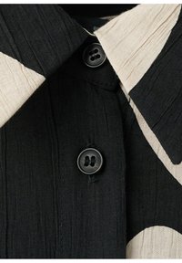 Černá látka s texturovanými svislými liniemi, zdobená dvěma černými knoflíky a límečkem s velkými béžovými a černými geometrickými vzory.