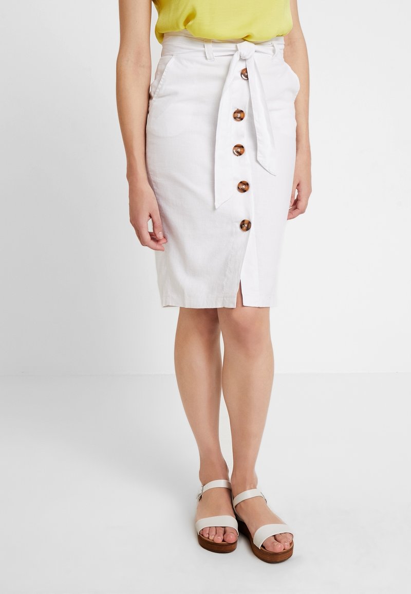 Femme portant une jupe blanche longueur genoux avec des boutons et une ceinture nouée, associée à des sandales beiges à brides et un top jaune.