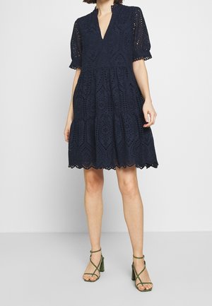 Femme portant une robe en dentelle ajourée bleu marine avec des manches bouffantes et des sandales à talons hauts à fines lanières vertes, debout sur un sol gris clair.