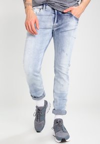 Světlé modré džínové kalhoty s efektem opotřebení a ohrnutými lemy, včetně vybledlého vzhledu a pětikapsového designu, doplněné šedými sportovními botami a bílými ponožkami.