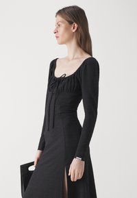 Black dress with long sleeves, square neckline, textured fabric, drawstring detail, and a thigh-high slit. Features HUGO label on the sleeve.