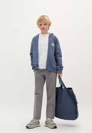 Felpa con zip blu navy, t-shirt bianca con grafica di un teschio, pantaloni grigi e sneakers in tonalità grigie. In mano tiene una grande borsa a tracolla blu. Design semplice.