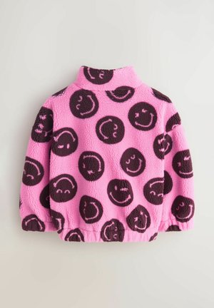 Vaaleanpunainen fleece-takki, jossa mustat hymyilevät kasvokuviot. Takissa on korkea kaulus ja joustavat hihansuut, jotka antavat istuvan muodon. Pehmeä materiaali.