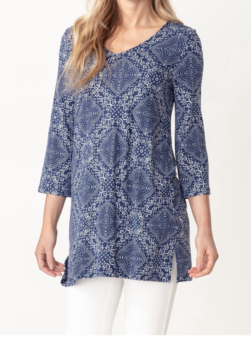 Femme portant une tunique bleue avec des motifs géométriques blancs et des manches trois-quarts, associée à un pantalon blanc, sur un fond uni.