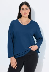 Navyblauwe lange mouwen V-hals shirt van gladde stof, met een relaxte pasvorm, zijsplitten en een ronde zoom. Draagt met zwarte broek.