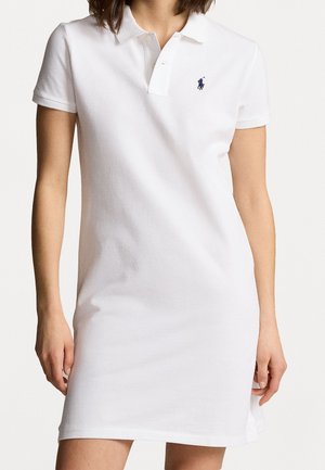 Kobieta w białej sukience polo z krótkim rękawem, z małym ciemnym haftowanym logo na lewej piersi.