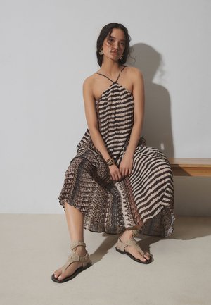 Femme en robe à col halter à motifs et sandales beige assise sur un banc contre un mur clair uni, les mains reposant entre les jambes.