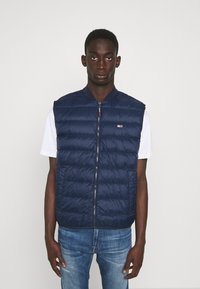Chaleco acolchado azul marino, con cremallera frontal, cuello acanalado y costuras horizontales, llevado sobre una camisa blanca. Estilo casual con aislamiento ligero.