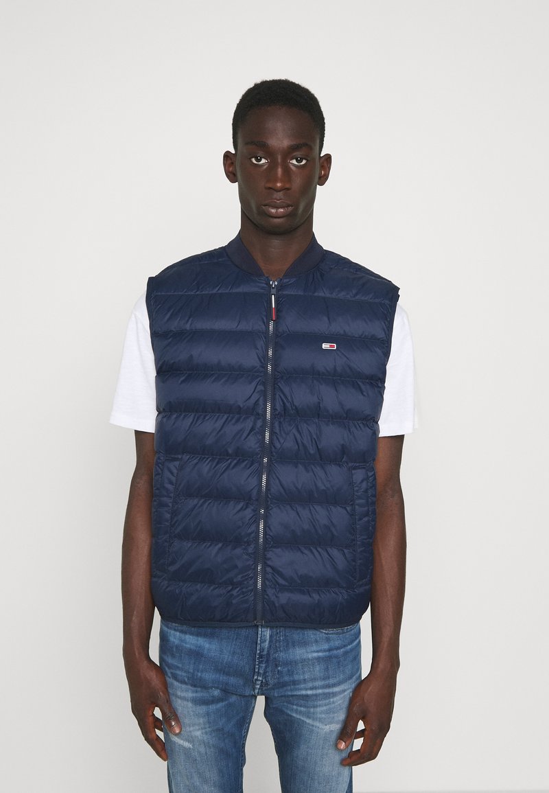 Chaleco acolchado azul marino, con cremallera frontal, cuello acanalado y costuras horizontales, llevado sobre una camisa blanca. Estilo casual con aislamiento ligero.