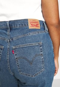 Niebieskie dżinsy Levi's z wysokim stanem, tradycyjnym wzorem pięciu kieszeni, brązową skórzaną naszywką z czerwonymi szczegółami graficznymi oraz subtelnym przeszyciem.