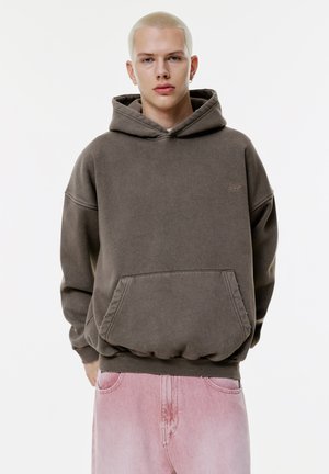 Haut à capuche brun surdimensionné avec une poche kangourou, un ourlet côtelé et un petit logo sur la poitrine, associé à un pantalon en denim rose clair.