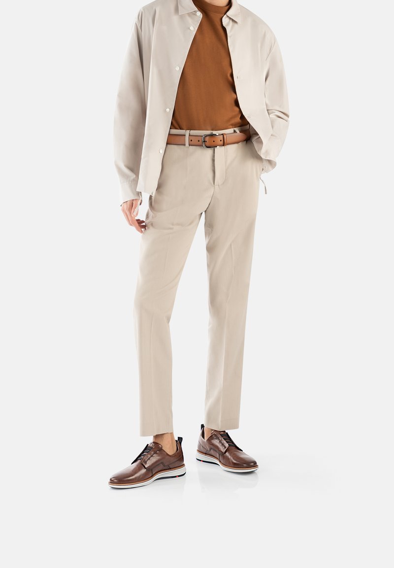 Beige jasje met knopen over een bruine shirt, gecombineerd met lichtbeige broeken en bruine leren schoenen met witte zolen.