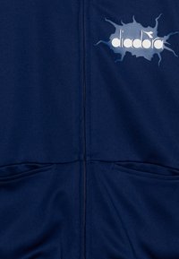 Σακάκι ανοιχτού μπλε χρώματος με φερμουάρ, δύο μπροστινές τσέπες και άσπρο λογότυπο "diadora" σε γραφικό σχέδιο  με ανοιχτό μπλε πιτσιλιές. Λείος υφαντικός ιστός.