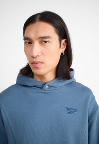 Blå hettegenser med kengurulomme, justerbar hals og brodert Reebok-logo i matchende farge. Glatt tekstur.
