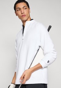 Λευκό πουλόβερ με φερμουάρ μέχρι το στήθος, κατασκευασμένο από υφασμάτινη υφή, με μαύρη λεπτομέρεια στο γιακά και λογότυπο στο μανίκι. Κρατώντας γκολφ κλαμπ στο χέρι.