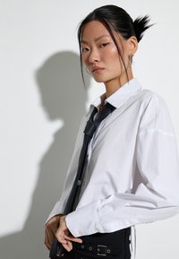 Camicia bianca con colletto, abbinata a una cravatta nera. Il modello indossa un indumento nero con un tessuto sembra strutturato, accessoriato con una cintura.