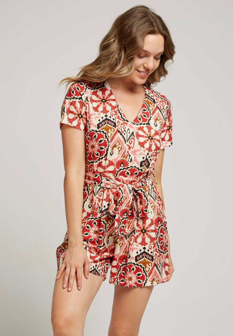 Rompers à imprimé floral en crème, avec des motifs rouges, noirs et orange. Manches courtes, décolleté en V et un détail de taille nouée avec des shorts.