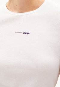 Bílý tričko s textem "Trvalá změna" vyšitým malým, smíšeným písmem na horním hrudníku.