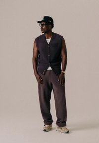 Vesta tricotată de culoare bleumarin cu un model texturat, cu nasturi pe față, purtată peste un top alb. Pantaloni de trening negri și adidași bej completează ținuta.