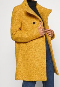 En person iført en livlig gul, struktureret frakke med høj krave står imod en lys baggrund og fremviser et stilfuldt og moderne look.