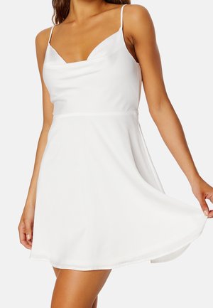 Kobieta ubrana w białą sukienkę bez rękawów z cienkimi ramiączkami i rozkloszowaną spódnicą, delikatnie trzymająca jedną stronę spódnicy.