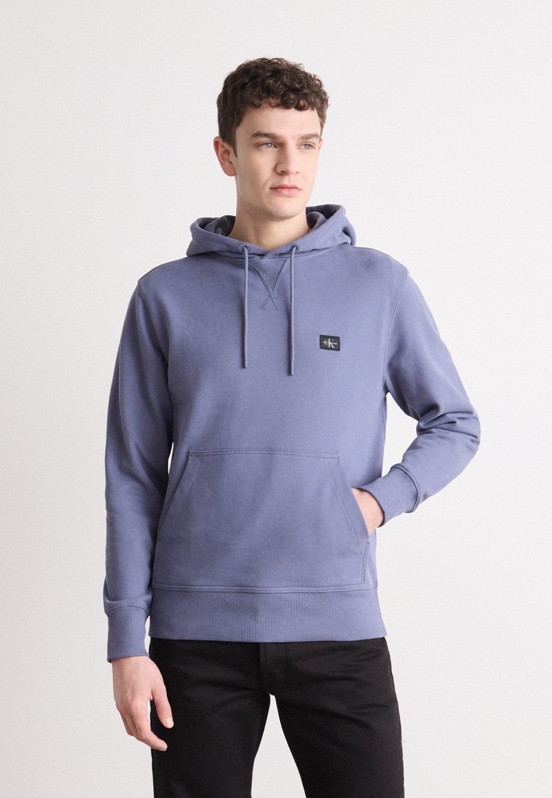 Felpa in cotone viola con tasca a marsupio, cordini regolabili e un piccolo patch con logo nero sul petto. Pantaloni neri.