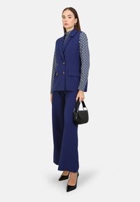 Marinblå skräddarsydd väst och vida byxor med en geometrisk mönstrad blus. Svart handväska med kort rem och spetsiga klackar.
