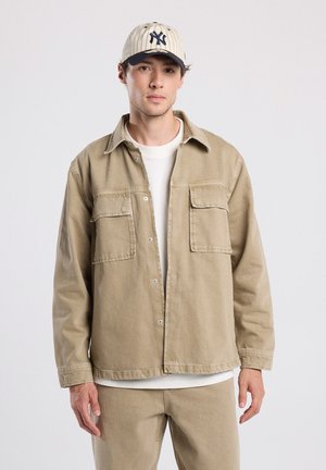 Beige denim jacket with two chest pockets, button closure, and long sleeves, paired with a white shirt and beige pants.