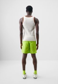 White ribbed tank top with red and yellow trim, paired with bright green shorts. Model wears green socks and white sneakers with blue accents.