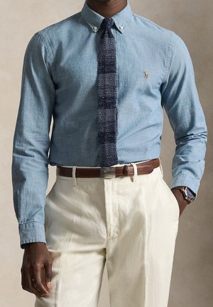 Homme portant une chemise bleu clair boutonnée avec une cravate texturée foncée, un pantalon beige, une ceinture en cuir marron et une montre-bracelet au poignet gauche.