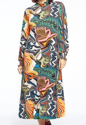 Kobieta w sukience z długim rękawem i wysokim kołnierzem o kolorowym, abstrakcyjnym wzorze z wirów w odcieniach pomarańczowego, zielonego, niebieskiego, różowego i czarnego.
