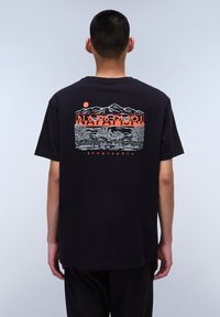 T-shirt noir en coton avec un imprimé graphique de montagnes et de soleil en orange et blanc au dos, avec le texte "NAPAPIJRI" au-dessus.