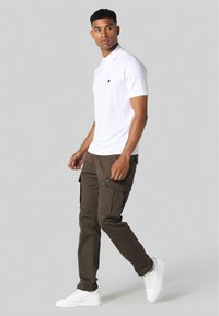 Hvit polo shirt med svart logo, kombineres med brune cargo bukser med sidelommer og en tapered passform, supplert med hvite sneakers.