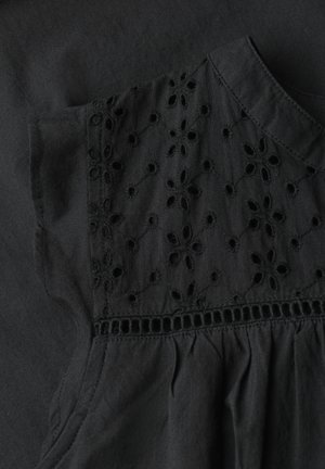 Detalje af sort stofkjole med blomsterudskåret broderi på den øvre brystdel, med opsamlet stof nedenunder og en rund halsudskæring.