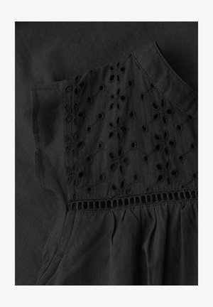 Detail eines schwarzen Stoffkleides mit floraler Lochstickerei auf der oberen Brustpartie, darunter geraffter Stoff und ein rundes Dekolleté.