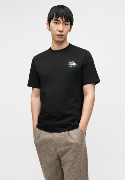 Μαύρο βαμβακερό T-shirt με στρογγυλή λαιμόκοψη, διακοσμημένο με πολύχρωμο κεντητό σχέδιο στο αριστερό στήθος. Το μοντέλο φοράει ταμπά υφασμάτινο παντελόνι με υφή.