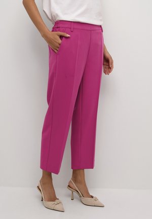 Vrouw in felle roze cropped broek met zijknopen en beige puntige hakken, staand tegen een plain witte muur.