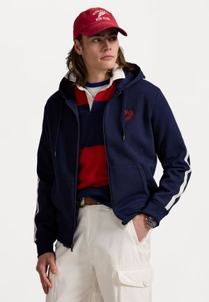 Felpa con zip blu marina con strisce bianche ai lati, logo rosso ricamato sul petto, indossata sopra una polo a righe rosse e blu marina, abbinata a pantaloni cargo bianchi.